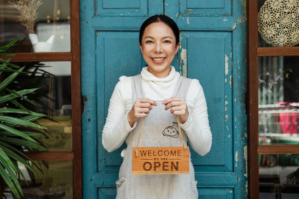 A smiling woman holding a sign saying "Welcome, we are open" while standing in front of her store front.